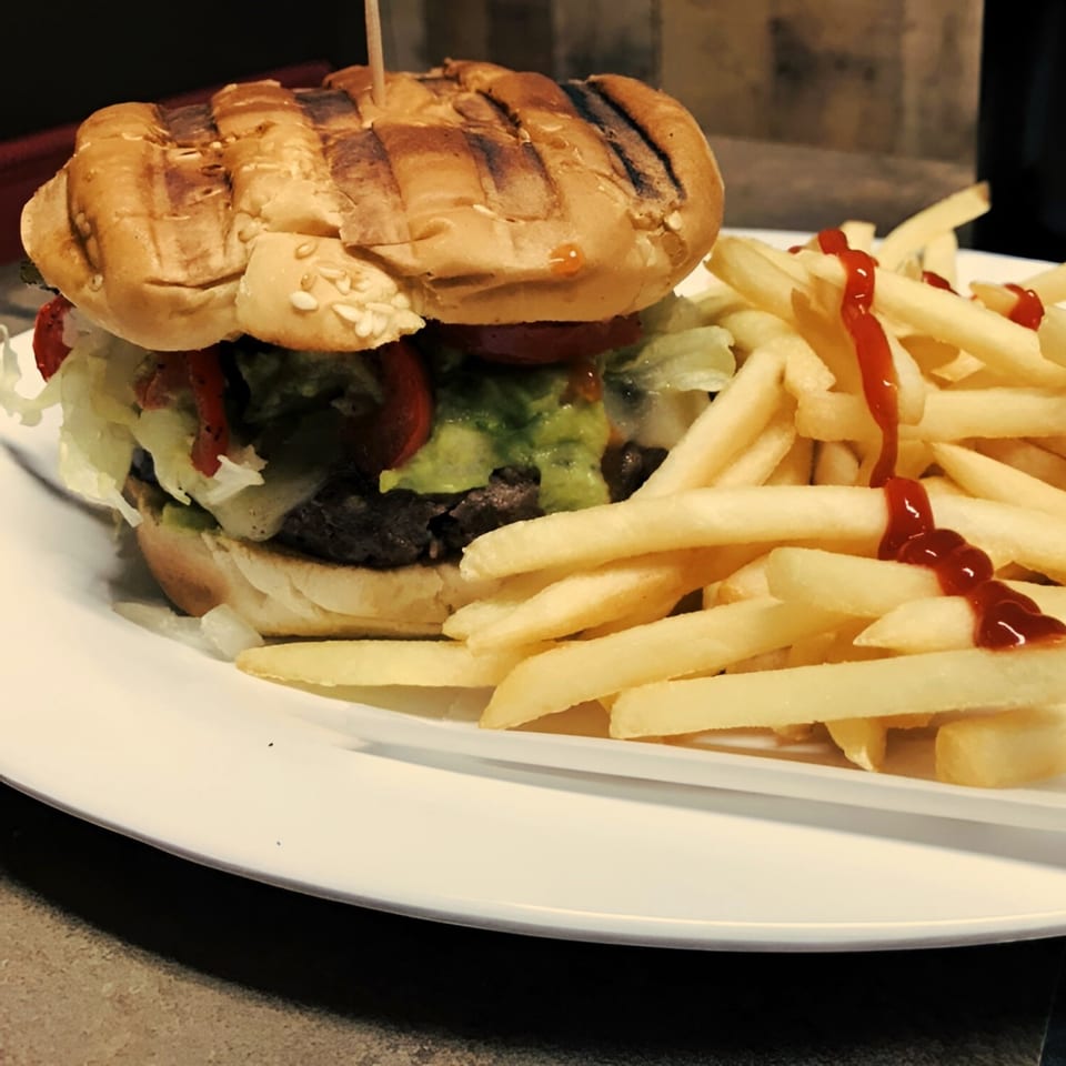 A grilled burger with guacamole, lettuce, and tomato, served with ketchup-drizzled fries.