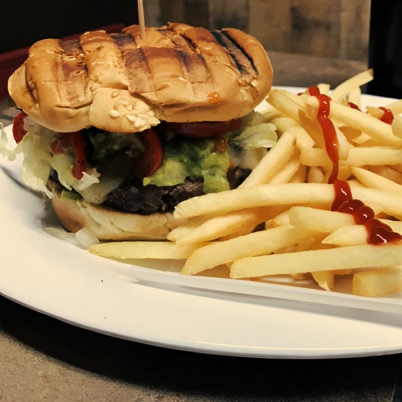A grilled burger with guacamole, lettuce, and tomato, served with ketchup-drizzled fries.