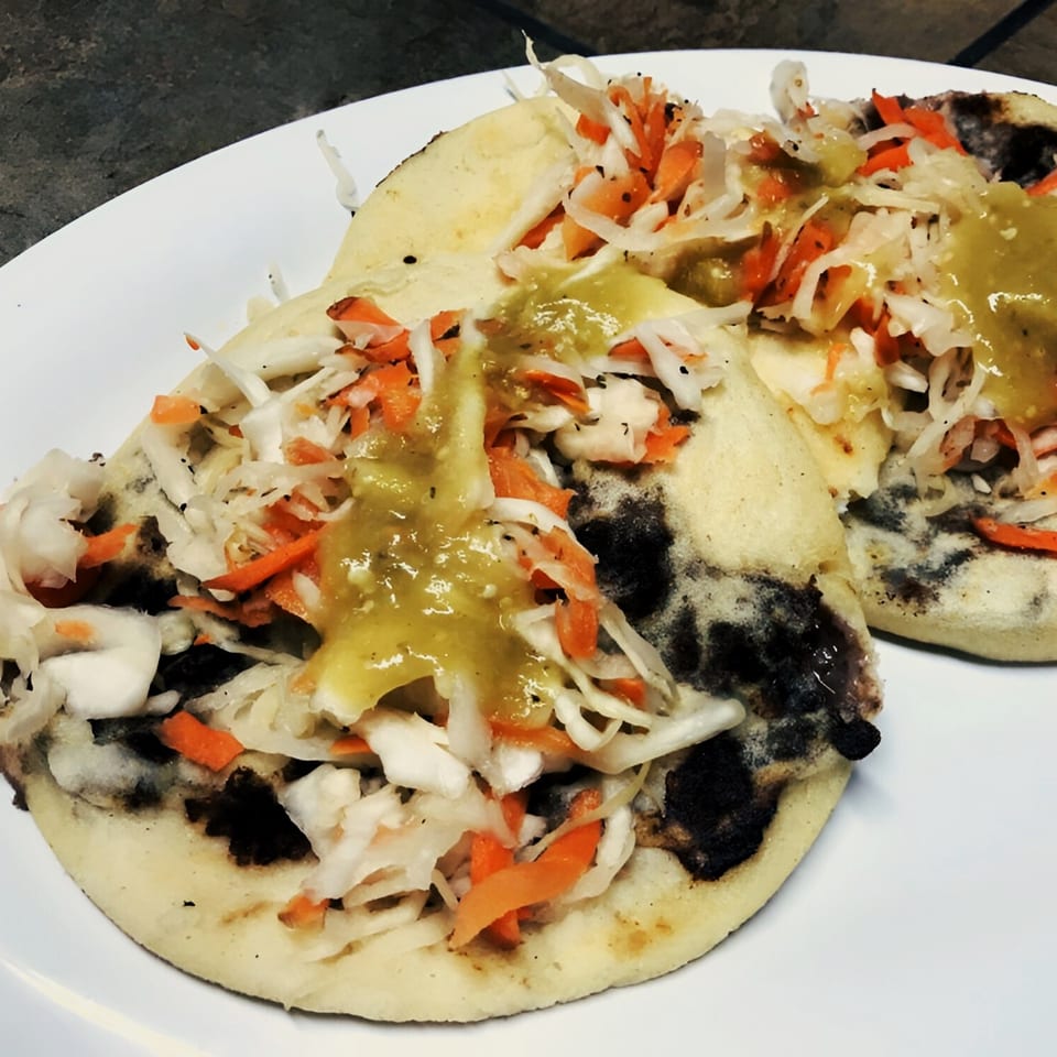 Two pupusas from El Vaquero, topped with curtido and green salsa on a white plate.