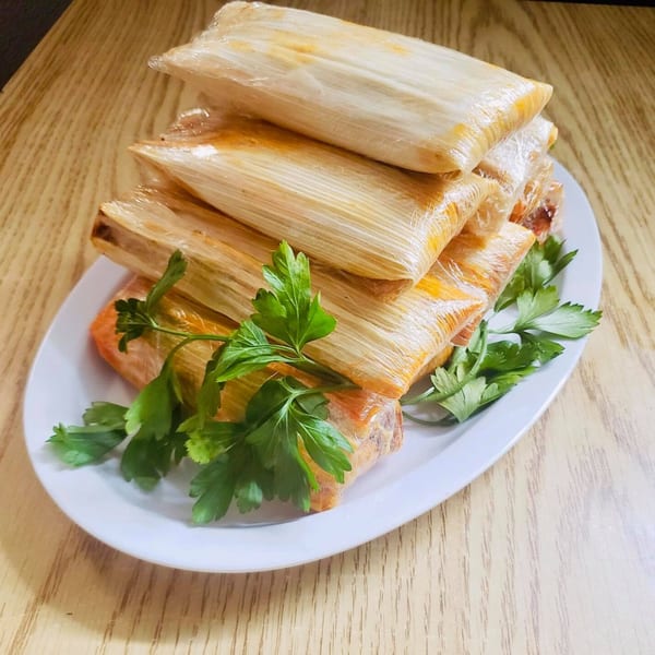 A white platter holds a stack of El Vaquero tamales wrapped in corn husks, garnished with parsley.
