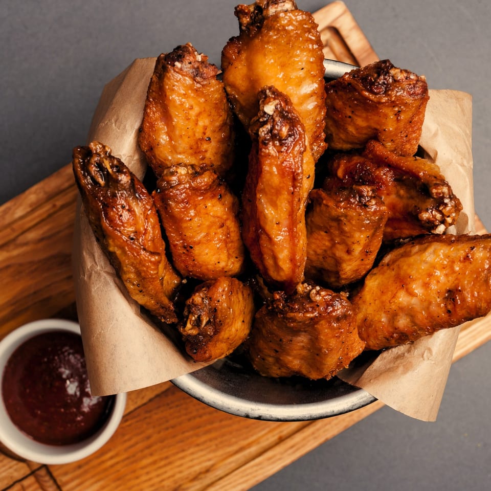 A metal bowl filled with crispy, glazed chicken wings and a side of dark dipping sauce.