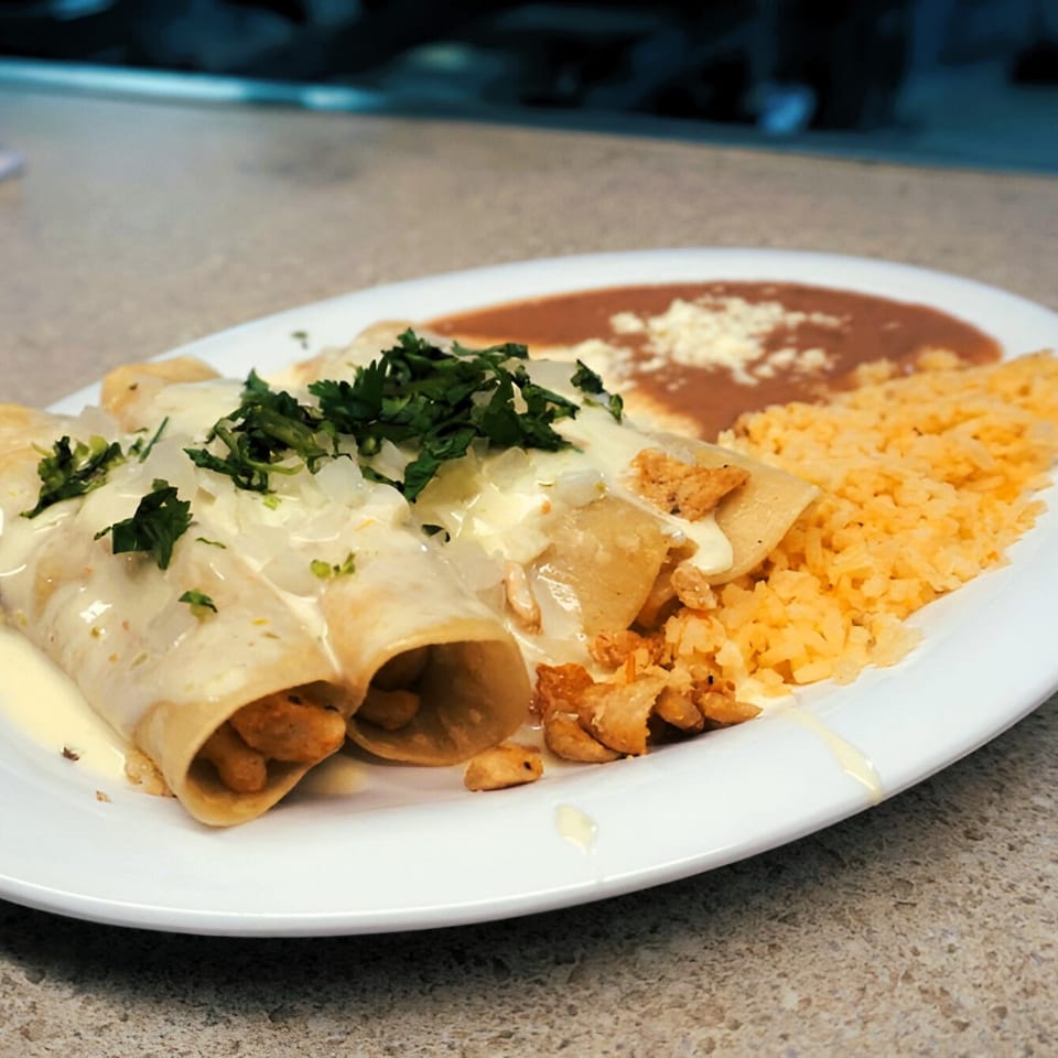 Chicken enchiladas with white sauce, cilantro, onions, rice, and refried beans on a white plate.