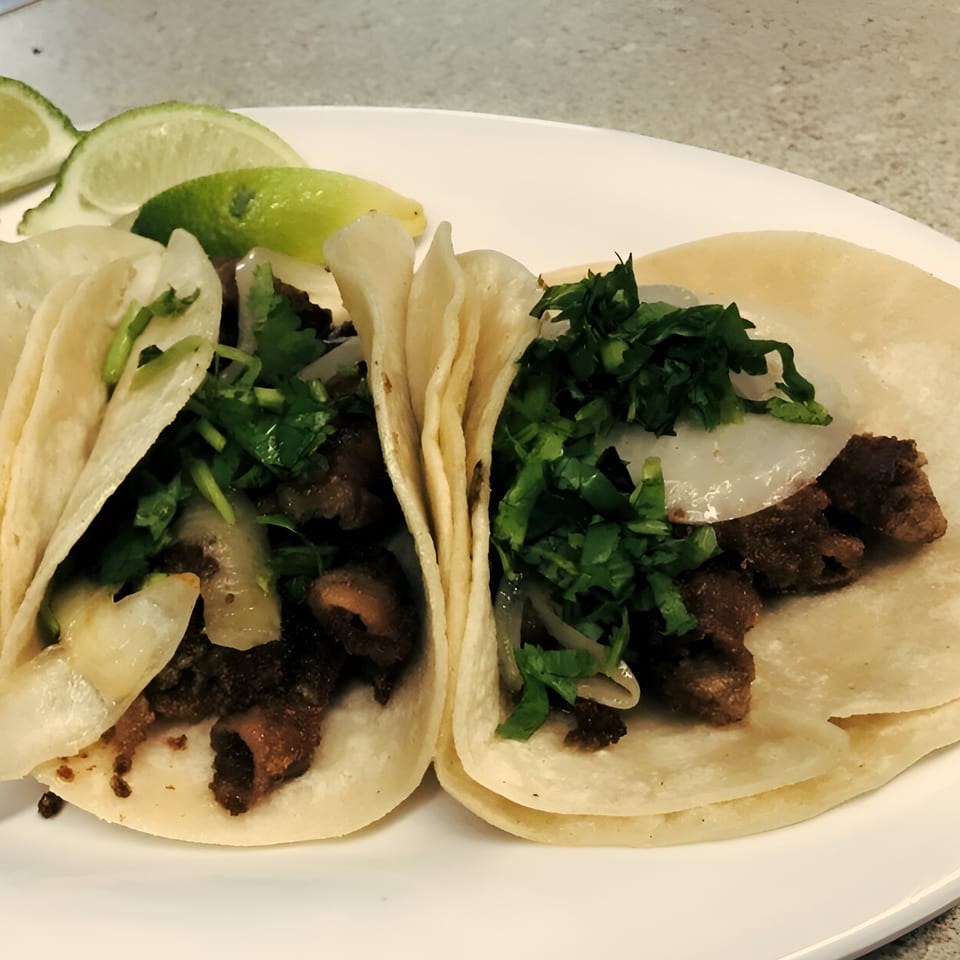 Two El Vaquero Mexican Restaurant tacos with meat, onions, cilantro, and lime wedges on a white plate.